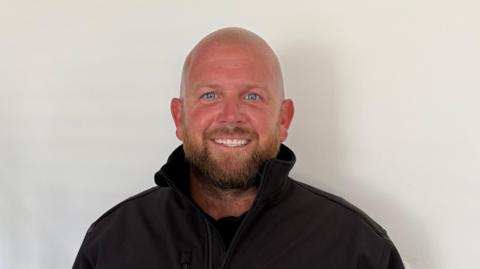 Jack McDermott, a man with a shaved head and short beard, smiles into the camera. He's wearing a black jacket with a high collar.