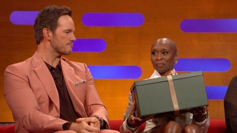 Cynthia Erivo holds a green gift box with a gold ribbon. Chris Pratt, to her right, looks at the box