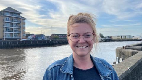 Georgia Shackleton, wearing a blue t-shirt, denim jacket, and glasses, has blonde hair. She is standing next to the River Yare in Great Yarmouth. There is a block of flats in the background.