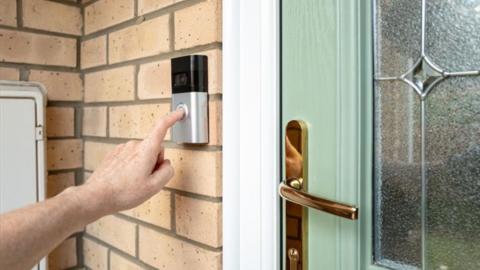 A hand reaches up to press a button on a smart doorbell at the front entrance of a home.