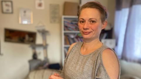 A woman wearing a grey jumper stands in her living room whilst looking into the camera.