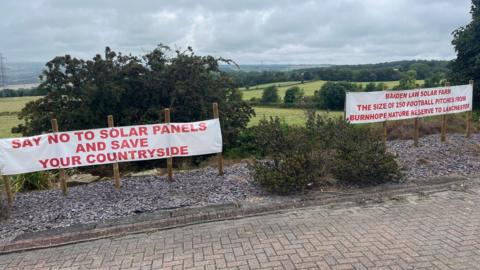 View of a landscape of fields with bushes and shrubs in the foreground and two posters, both objecting to plans for solar panels