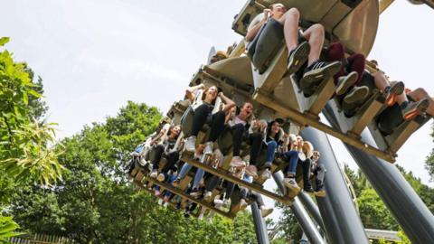 A group of visitors are on a ride in Alton Towes sitting with their feet dangling from the ride. There are green trees in the background and the people are laughing and smiling. The picture is taken from the ground.
