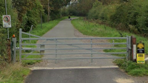 A large silver gate blocks the entrance to the bridleway, which is long and flanked by trees and bushes.