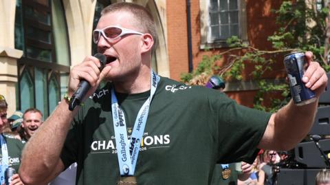 Alex Coles speaking into a microphone wearing sunglasses, a green T-shirt and a medal round his neck. He is holding a can of beer in his other hand.