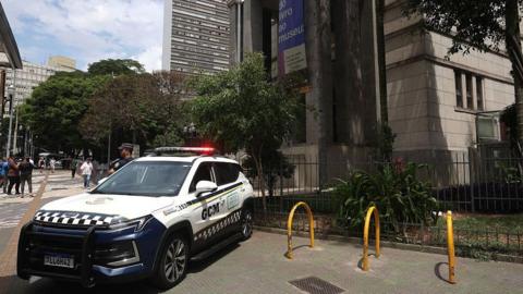 A police car with its lights on is seen parked on a pavement outside the library - a large grey building - as pedestrians walk in the background. A police officer with his arms crossed is seen stood next to the car