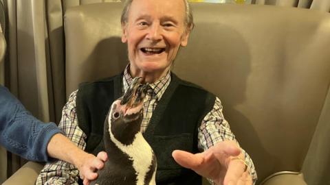 An older man smiles as he holds a penguin at a care home