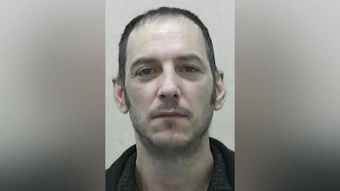 Mugshot of Mark Harvey. He has pale, pasty skin, scruffy short dark hair and a faint wispy dark beard