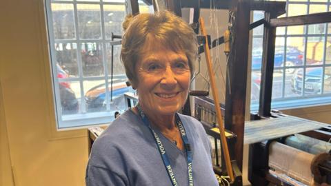 A woman with short brown hair is pictured wearing a blue knitted jumper with a blue lanyard with wording that reads "volunteer" in white capital letters. She is stood in front of a window and also next to a wooden weaving loom. 
