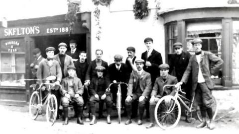 An old black and white photo of men in flat caps posing with bicycles. 