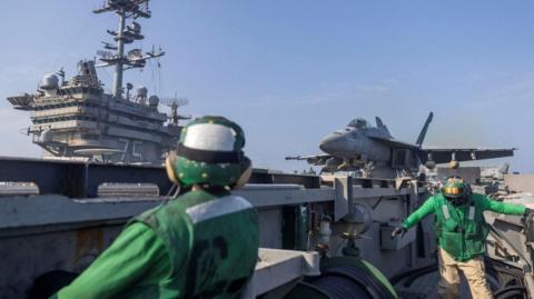 Two US military members in bright green jackets stand on an aircraft carrier with a jet behind them