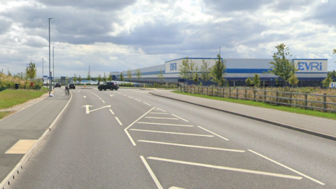Street View image of a new-looking road with an industrial estate on the right. There is a large Evri dispatch factory. A car is pulling out of the road on the right.