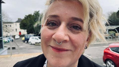 The image shows an outdoor setting with a paved area in the foreground and a car park in the background. The person in the foreground is wearing a black jacket and a white lanyard. Their hair is light-coloured and styled in loose waves. Several cars are parked, including a white car, a yellow car, and a red car. Behind the car park, there are trees and a light-coloured building with multiple windows and a bright red door.