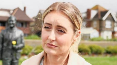 A blonde woman standing, looking past the camera. There is a metal statue over her right shoulder holding a yellow flower.