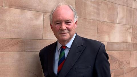 A man in a black suit jacket, navy, red and green striped tie and white shirt standing outside against a brick wall. he has white receding hair.