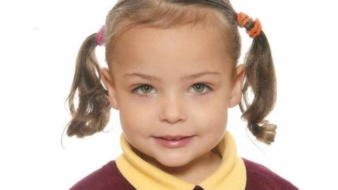 Poppy has hazel eyes and is wearing her blonde hair in two side pigtails. She is wearing a burgundy jumper over a yellow polo shirt.
