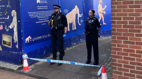 Two police officers stand in front of a police cordon of blue and white tape.