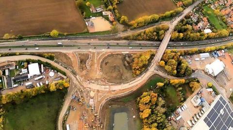 A birds-eye-view image of the roadworks. It shows a new junction that is being built above two busymain roads. To the left there is a large building with solar panels.