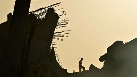Person in silhouette against collapsed building. The background is a warm yellow while the remains of the building and the person are dark.