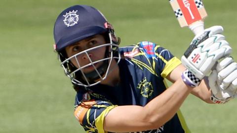 Maia Bouchier wearing a blue and yellow Hampshire shirt and blue helmet swings her bat