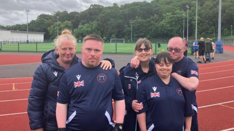 Two able bodies women - one blonde and on with brown hair - stand on a athletics track with two men and one woman who have Down Syndrome. They are wearing dark blue Team GB kit