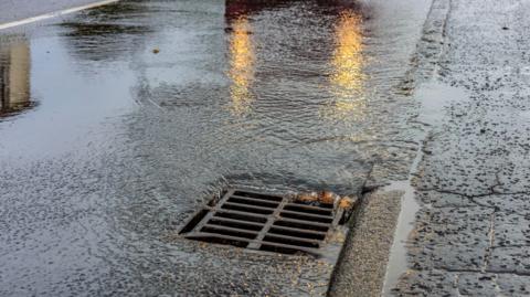 A stock image of a drain cover with a car's headlights illuminating the road. Water is pouring into the drain.