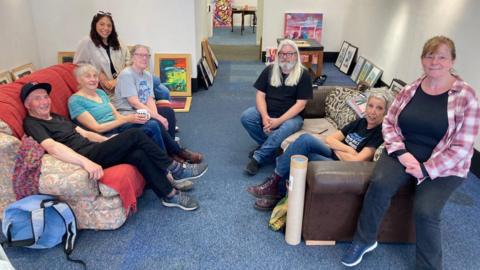 Seven people sitting on two sofas in a white-walled room with paintings on the floor leaning on the walls.
(Left to right) Barry Greenwood, Caro Blount-Shah, Ann Driver, Lauren Kelly, Ken Woods, Cath Muldowney, Pauline Cooke