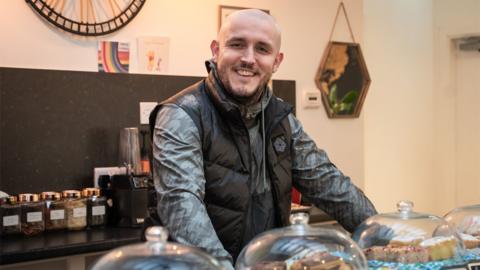 Former prisoner Lee Jack, wearing a black coat, smiles while looking at the camera - he's standing behind a cafe counter with cakes and biscuits in glass jars