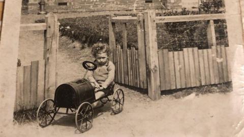 A black and white photo of a young boy sitting in an old-fashioned small car toy. Behind him is a wooden fence with an open gate with a front garden and a brick house in the background.