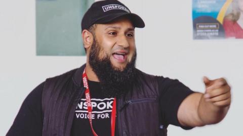 Atiq Rehman making a speech wearing a cap with the word "Unspoken"