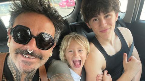 A man with black sunglasses on in the back seat of a car with two boys.