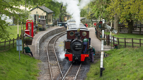 An historic steam train waiting at the platform at Whipsnade Zoo railway. It is surrounded by mature trees and grassy embankments.