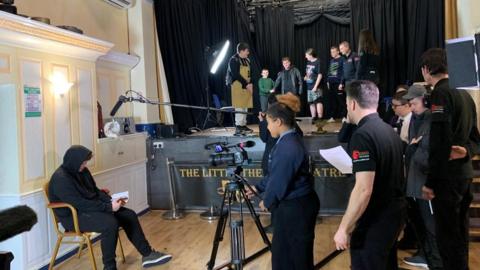 A TV crew filming a man sitting on a chair in a room. There is a camera, lights and mics surrounding the man. There is a stage behind the crew with seven people standing on it. 