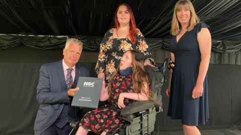 Group portrait on a stage with four people, one of them is in a powered wheelchair holding a National Star College Record of Achievement folder. Another person kneels beside the wheelchair presenting the folder, while two others stand behind. The wheelchair user wearing a floral-patterned dress and the presenter dressed in a suit and tie.
