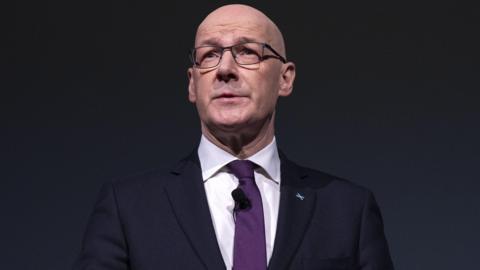 John Swinney wearing a dark suit over a white shirt and purple tie standing against a black background with his hands interlocked in front of him.