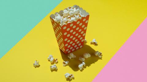 A polka dot (white dots on red background) carton of popcorn on bright background of three coloured diagonal stripes which are turquoise, yellow and pink. 