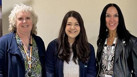 Three women standing together in front of a wall. The woman on the left has white blonde curly hair, a black, white and green dress and a navy blue cardigan. In the middle is Leanne, who has long brown hair, a white top and navy zip hoodie. On the right the final woman has long black hair, a zebra print top and a black leather jacket on. 