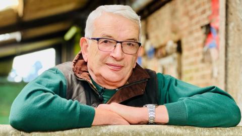 Michael Read leans on a wooden gate at his farm. He has white hair and wears glasses and a green jacket. Behind him stands a barn with a red-brick wall.