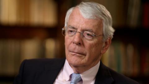 Sir John Major, wearing a dark suit, with a blue tie and white shirt, in a reflective mood
