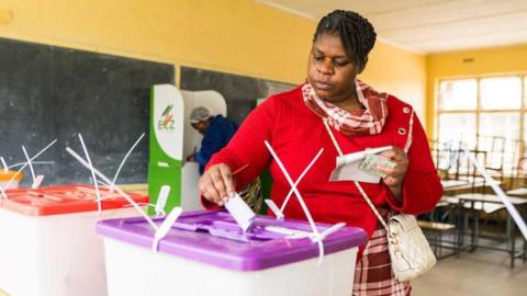 Inside a school classroom in Lusaka, Zambia, a woman wearing a red top places a ballot paper in a plastic ballot box with a purple lid. Some desks can be seen behind her.