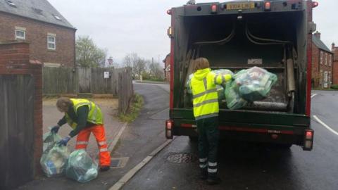 A man with a hi-vis vest picking up rubbish bags and another man with a hi-vis vest putting bags into a bin lorry. It is a residential street with houses.