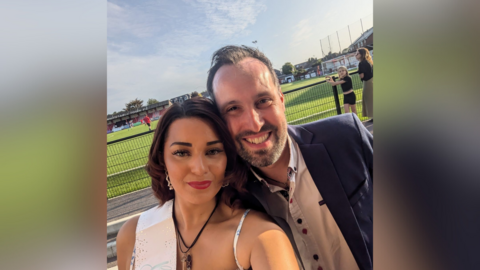 Charlotte Rickwood and Jordan Collins pictured at Charlotte's hen do. She is wearing a white sash which says 'Bride Squad'. Jordan is wearing a navy suit jacket and a white shirt. They are both stood in front of a football pitch where a game is ongoing.