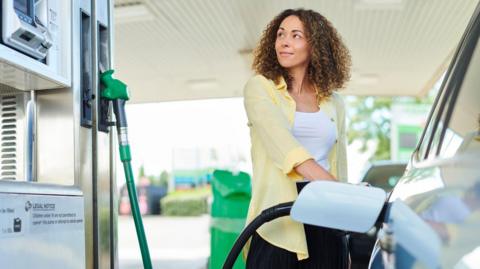 A woman wearing a white top and yellow shirt looks at a pump while filling up her car.