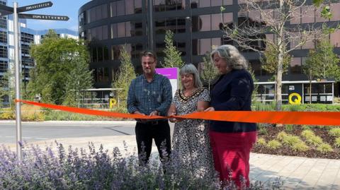 Three people stand behind a large red ribbon.  Councillor Chris Watts, mayor Fay Howard and MP Heidi Alexander are getting ready to cut the ribbon. They are standing in a public area, with freshly planted trees and flowers, clean pavements and fresh tarmac. A large building with glass windows is behind