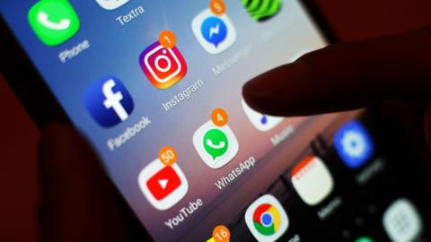 Stock image of a thumb scrolling through a mobile phone. There are several icons on the smart phone, including Whatsapp, YouTube and Instagram.