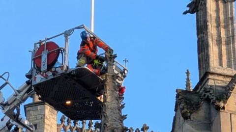A person wearing a blue helmet and orange clothing is positioned on an aerial platform above Hull Minster. They are cutting a decorative stone finial on the roof of the building.