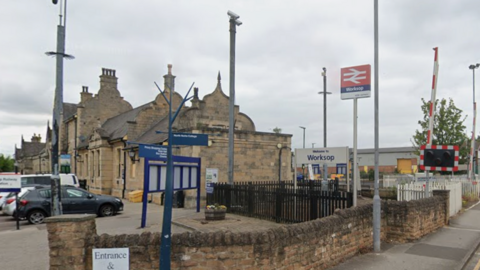 An image of a Victoran brick railway statiom with cars parked outside and a level crossing barrier to the right