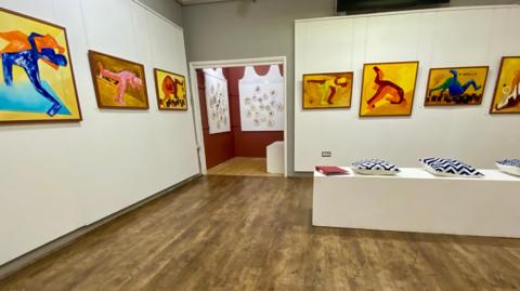 Gallery with colourful paintings on a white wall depicting abstract wrestlers. They are entwined in various positions.