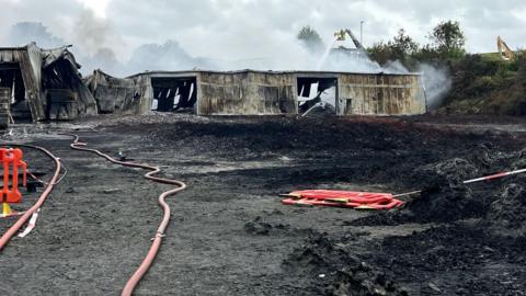 A burnt out building, a large fire hose, red plastic barriers on the ground, smoke in the air, and water being poured on a metal building that is crumpled and collapsed