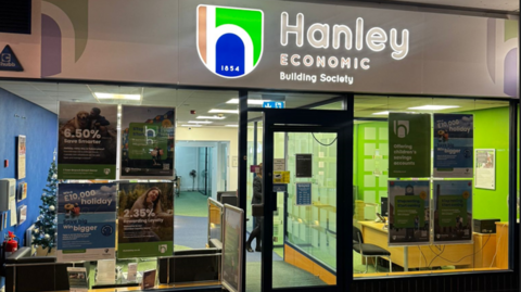 The front of a retail unit, which is a framed in blue, and has posters in the windows advertising financial products.